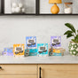 Boxes of Molly's Suds Laundry Detergent Sheets in Citrus Grove, Peppermint, Ocean Mist, and Lavender scents are displayed on a kitchen counter with a plant, emphasizing their plastic-free packaging.