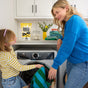 A woman and a young girl smile as they load laundry into a front-loading washer in a bright room. On the counter are detergent, Molly's Suds Oxygen Whitener Bleach Alternative, and a vase of yellow flowers.
