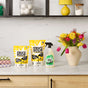 A kitchen counter displays Molly’s Suds Oxygen Whitener Bleach Alternative, a spray bottle, colorful folded towels, and a vase of yellow, pink, and red flowers near shelves with jars and laundry supplies.