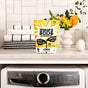 A package of Molly's Suds Oxygen Whitener sits on a white shelf above a washing machine, with folded towels and a vase of yellow and white flowers in the background.