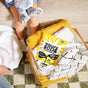 A child with a stuffed animal sits on a couch beside a wire laundry basket of yellow and white towels and a bag of Molly's Suds Oxygen Whitener, on a green and white checkered rug.
