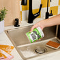A hand pours Molly's Suds Sink SCRUB™ Sink Cleaner into a kitchen sink. Nearby are a brown sponge, soap dispenser, small potted plant, and folded yellow and pink towel—everything for a gentle scrub.