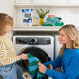 A woman and a young girl smile as they load striped laundry into a front-loading washer. Molly's Suds Super Powder Laundry Detergent with Enzymes for sensitive skin sits on the counter above, making laundry time enjoyable together.