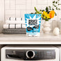 A bag of Molly's Suds Laundry Detergent Powder rests on a laundry room counter beside folded towels, a checkered cloth, and yellow flowers. Below is a front-loading Electrolux washer. Gentle on sensitive skin.