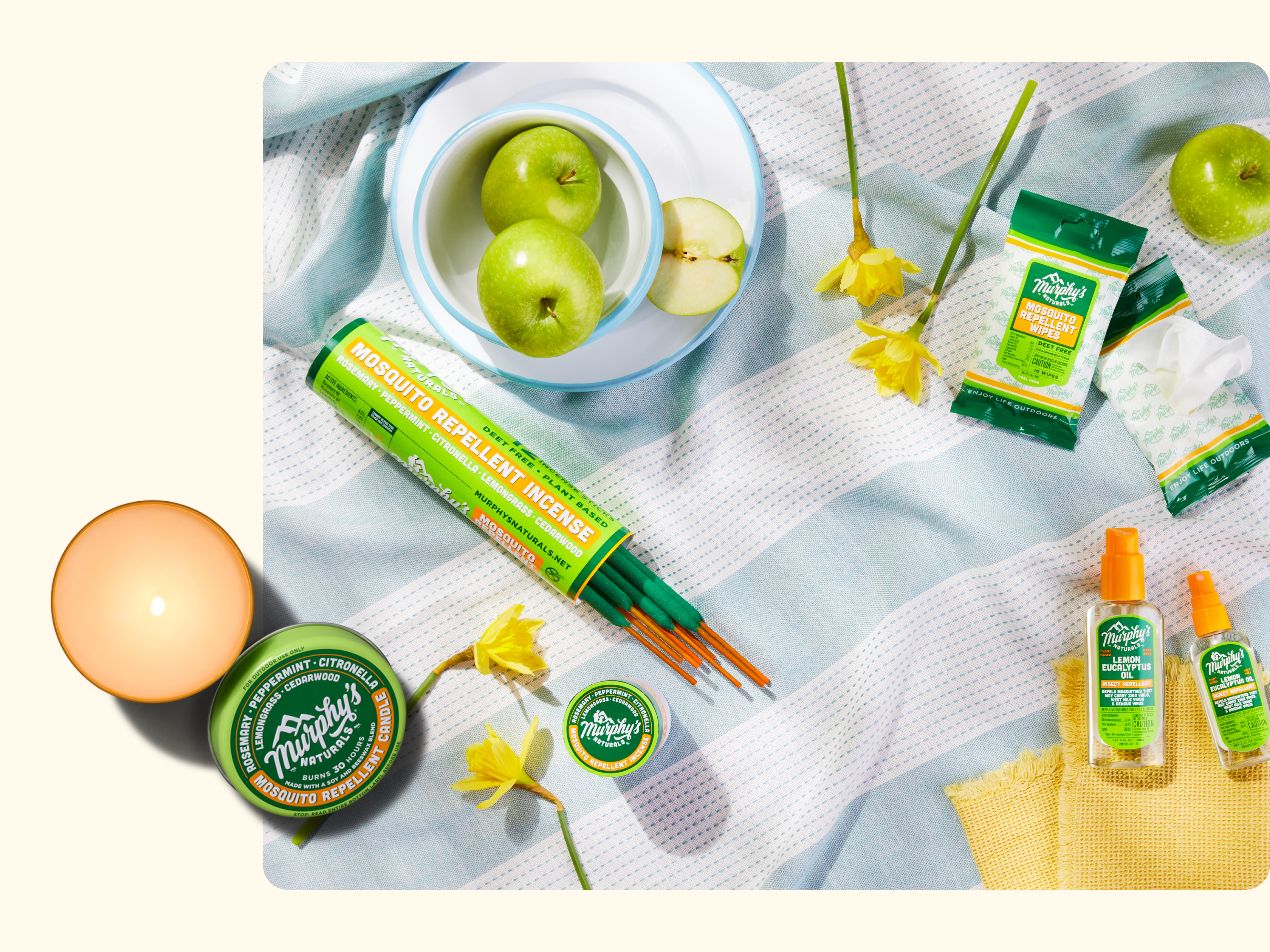 A flat lay of Murphys Naturals mosquito repellent products on a striped cloth, surrounded by green apples, yellow flowers, a bowl, a candle, and a yellow mesh bag.