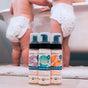 Three bottles of Motherlove Baby & All Castile Soap—lavender, grapefruit, and unscented—sit on the floor in front of two toddlers in diapers by a white bathtub.