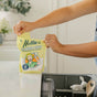 Someone holds a yellow Nellie's Dishwasher Powder Pouch above an open dishwasher with clean dishes. The pouch features a cartoon woman, notes it’s phosphate-free, septic safe, and provides 50 loads per package.