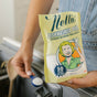 A person holds a blue scoop of Nellie's Dishwasher Powder Pouch, a phosphate-free white powder, over a dishwasher dispenser. The yellow pouch displays the Nellie's brand and features a retro illustration of a smiling woman.