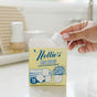 A hand takes a Nellie's Dish Cubes, 16 Load, from its open box on a white kitchen counter. The box highlights highly concentrated cleaning power, plastic-free packaging, and enough cubes for 16 dishwasher loads.