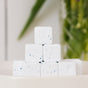 A small pyramid stack of Nellie's Dish Cubes, 16 Load—highly concentrated cleaning tablets with blue specks—displayed on a white surface with a blurred green and floral background.