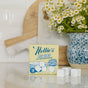 A box of Nellie's Dish Cubes, 16 Load, sits on a white counter beside a blue and white vase of small white flowers. Three concentrated cleaning cubes are in front, with a wooden cutting board in the background.