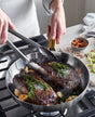 Using tongs, someone cooks two rosemary and garlic-seasoned steaks in a GreenPan Reserve Stainless Steel Cookware Set on the stove, with red pepper flakes and sliced vegetables in bowls nearby.