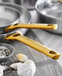 Two GreenPan Reserve Stainless Steel Cookware pans with gold-colored handles sit on a wooden surface, next to a bowl of salt and pepper mix and two garlic bulbs.