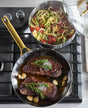 Two pans from the GreenPan Reserve Stainless Steel Cookware Set: one with seared steaks, rosemary, and garlic; the other with zucchini noodles, cherry tomatoes, and herbs—both cooked in PFAS-free cookware.