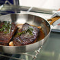 Using an oven mitt, a person removes the GreenPan Reserve Stainless Steel Open Fry Pan from the oven, revealing two seared steaks with rosemary, for restaurant-quality cooking at home.
