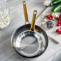 Two GreenPan Reserve Stainless Steel Open Fry Pans with gold handles rest on a light gray kitchen counter near a cutting board topped with veggies, garlic, spices, and a jar of white beans. Tri-ply stainless steel ensures even heating.