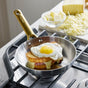 A fried egg tops a grilled ham and cheese sandwich in the GreenPan Reserve Stainless Steel Open Fry Pan Set, with melted cheese oozing out. Shredded cheese and a bowl of butter sit on a nearby cutting board.