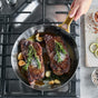 A person cooks two seared steaks with rosemary and garlic in a GreenPan Reserve Stainless Steel Open Fry Pan Set on the stovetop, with seasonings, lemon slices, and a small bowl arranged on a nearby cutting board.