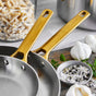 The GreenPan Reserve Stainless Steel Open Fry Pan Set features two tri-ply pans with gold handles, stacked on a kitchen counter beside pasta, garlic bulbs, a bowl of salt and pepper mix, and fresh herbs.