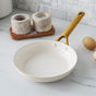 The GreenPan Reserve Pro Open Frypan, a white ceramic nonstick pan with a gold handle, sits on a marble countertop. Beside it are two brown eggs and two stone salt cellars on a wooden tray, against a tiled backsplash.