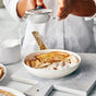 A person dusts powdered sugar over a baked apple dessert in a white dish using a small sieve. Nearby, a GreenPan Reserve Pro Open Frypan and a glass container of powdered sugar with a gold-handled spoon are visible.