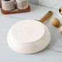 The GreenPan Reserve Pro Open Frypan, a white ceramic nonstick pan with a gold handle, sits upside down on a marble countertop near brown eggs and a wooden tray. Its engraved base highlights its toxin-free cooking design.