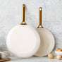 Two white GreenPan Reserve Pro Frypans with gold handles lean against a tiled wall. Nearby, brown eggs and a speckled bowl sit on the counter—perfect for PFAS-free cooking with this ceramic nonstick set.