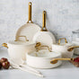The GreenPan Reserve Pro Cookware Set, featuring PFAS-free cream-colored ceramic nonstick pots and pans with gold handles and glass lids, is displayed on a marble countertop beside utensils, apples, and flour jars against a white tile backsplash.