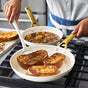 Wearing a blue apron, a person flips French toast in a white ceramic nonstick Reserve Pro skillet by GreenPan, while another PFAS free pan from the Reserve Pro Cookware Set simmers vegetable stew on the stove nearby.