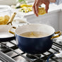 A hand drops pasta into a large navy blue GreenPan Reserve Pro Cookware Set pot with gold handles, filled with boiling water on a stovetop in a bright kitchen.