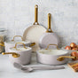 The GreenPan Reserve Pro Cookware Set, a PFAS-free light gray ceramic nonstick collection with gold handles—including three lidded pots and two frying pans—rests on a marble kitchen countertop near utensils and onions.