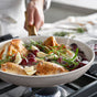 A person cooks chicken, garlic cloves, grapes, and rosemary in a frying pan on the stove. Steam rises as they hold the pan, demonstrating the safety and ease of GreenPan’s PFAS-free Reserve Pro Cookware Set with ceramic nonstick coating.