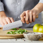 A person chops green herbs on a wooden cutting board with a GreenPan Elite Light Pakka Wood Cutlery knife. A bowl of salt and a yellow tomato rest nearby on the counter.