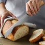 Someone slices a loaf of rustic bread on a wooden surface with a GreenPan Elite Light Pakka Wood Cutlery Knife, two slices already cut, while holding the bread steady with a cloth.