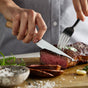 A person slices a medium-rare steak on a wooden board using the GreenPan Elite Light Pakka Wood Steak Knife Boxed Set, with herbs, salt, and garlic beside the meat.