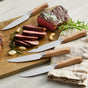 The GreenPan Elite Light Pakka Wood Steak Knife Boxed Set features four ergonomic steak knives with wooden handles, beautifully arranged on a cutting board beside sliced steak, garlic, rosemary, and a beige napkin—ideal for any meal.