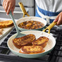 Wearing a white shirt and apron, a person cooks three slices of French toast in a GreenPan Reserve Pro Frypan Set on the stove, with another frypan simmering fruit compote nearby.