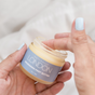 A hand with light blue nail polish dips a finger into a jar of Naturally London Nourishing Foot Butter, enriched with Cupuaçu, mango, and avocado butters, against a soft neutral background.
