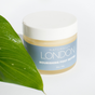 A jar of Naturally London Nourishing Foot Butter with a white lid sits on a white surface beside a green leaf. Its light blue label highlights this moisturizing foot cream made with Cupuaçu, Mango, and Avocado butters.