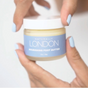 A person with light blue painted nails holds a small jar of Naturally London Nourishing Foot Butter against a light background.