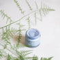 A jar of Naturally London Nourishing Foot Butter in light blue rests on a white surface, artfully surrounded by delicate green fern leaves.