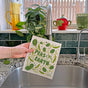 A hand holds The Neighborgoods Dish Towel + Sponge Cloth – Peas on Earth, featuring green leaves and text, over a kitchen sink. In the background, plants, a red teapot, and a green mug are seen on the windowsill.