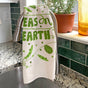 A Dish Towel + Sponge Cloth - Peas on Earth by The Neighborgoods hangs over a faucet, with a potted plant and eco-friendly yellow bottle resting on the windowsill above the granite countertop.