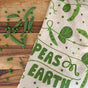 The Neighborgoods Dish Towel + Sponge Cloth - Peas on Earth, featuring pea plant illustrations and the phrase PEAS ON EARTH, rests on a wooden surface surrounded by fresh peas and snap pea pods.