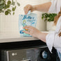 A person in a white shirt with rings opens a blue bag of Nellie's Laundry Soda on top of an LG washing machine, with plants visible in the background.