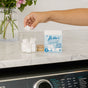 A hand places a white Nellie’s Laundry Cubes, 16 Load, into a glass jar beside an open box on a marble countertop above a washing machine, with flowers in a vase in the background.