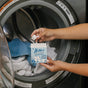 A person holds a box of Nellie's Laundry Cubes, 16 Load, ready to drop a concentrated, PVA-free cube into an open washing machine with white and blue towels.