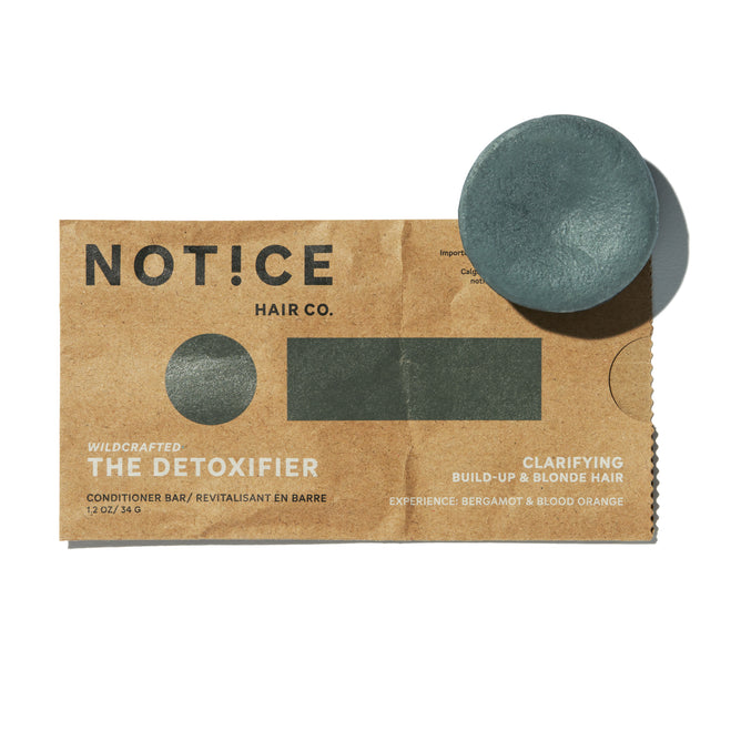 A round green The Detoxifier Conditioner bar by NOTICE Hair Co sits on a brown package, offering plastic-free care for buildup and blonde hair with activated charcoal, bergamot, and blood orange.