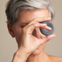 A person with short gray hair holds NOTICE Hair Co’s The Detoxifier Conditioner bar—a round, dark gray clarifying bar—over one eye, while the other remains visible. The background is a neutral beige.