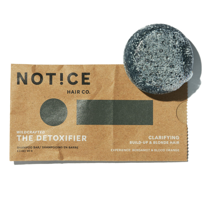 A round, speckled grey The Detoxifier Shampoo bar by NOTICE Hair Co sits on a brown paper label. It’s infused with activated charcoal and features bergamot & blood orange scents for clarifying, build-up removal, and blonde hair care.